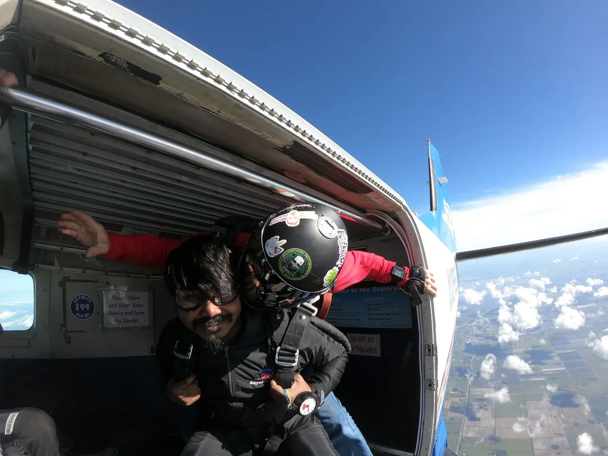Bheeshma smiling in the open door of the plane, strapped to a tandem instructor, fields and clouds far below